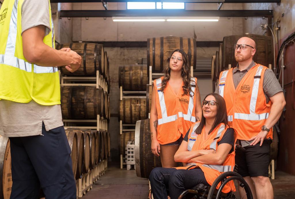 Three people - two standing, one in a wheelchair , wearing orange safety vests and glasses facing a person in a yellow safety vest